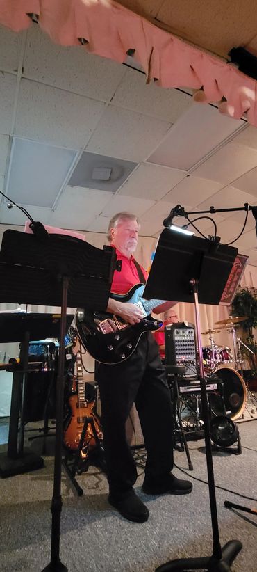 Man playing electric guitar on stage with music stands and drums in the background.