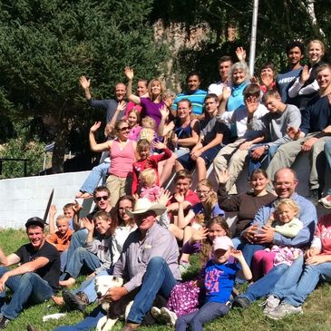 A large group of people smiling and waving outdoors on a sunny day.