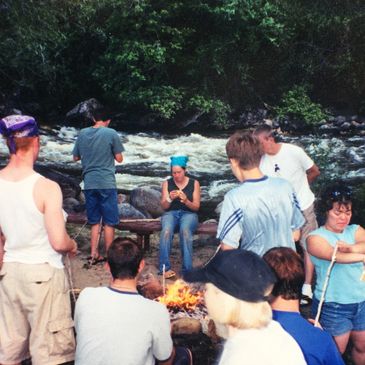 Group of people gathered around a campfire near a river in a forested area.