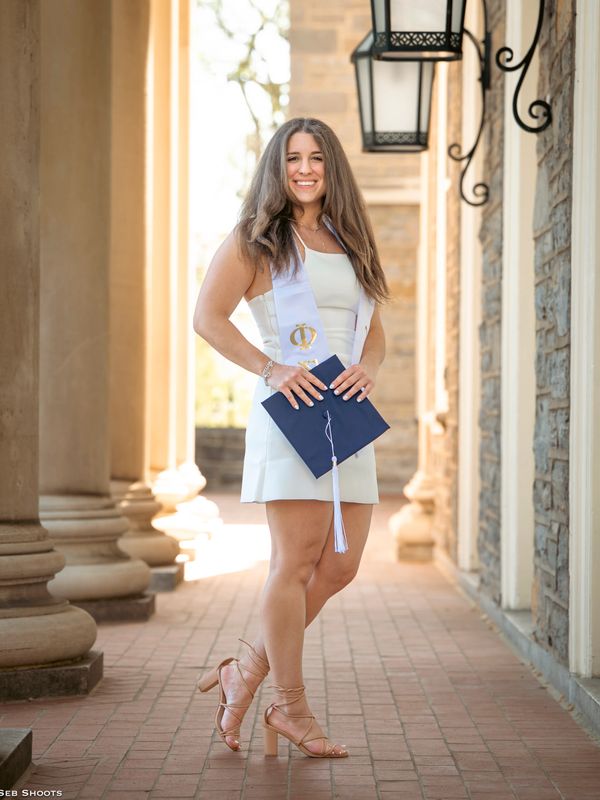 Young woman in white dress holding graduation cap, smiling in a bright corridor.