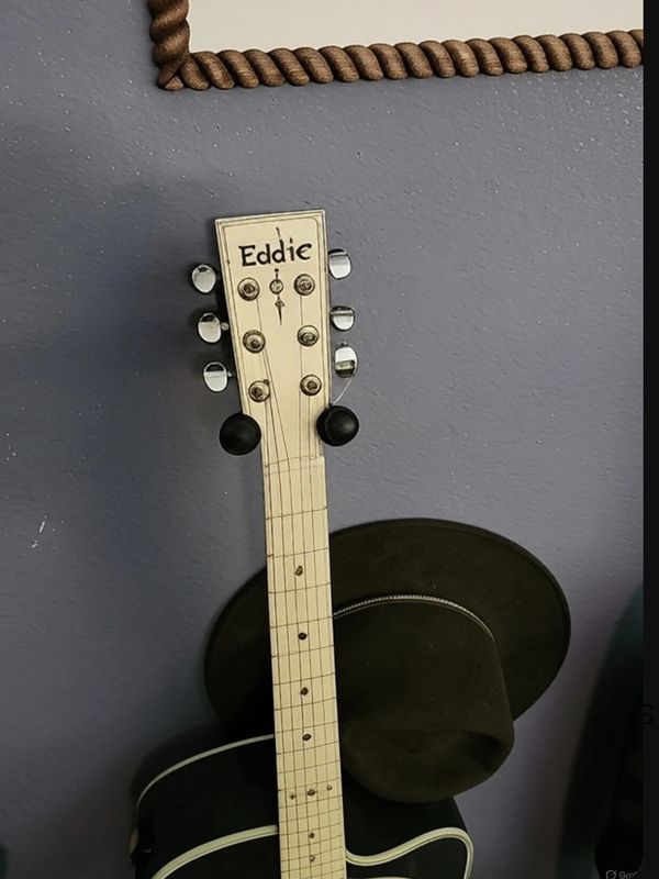 White guitar headstock labeled 'Eddie' leaning against a wall with a black hat.