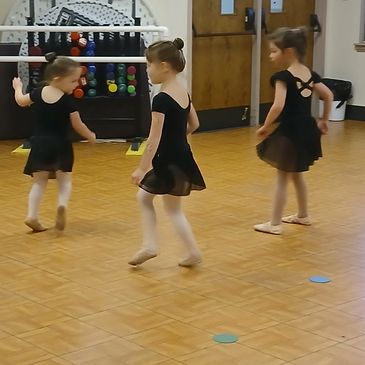 Three young girls in black ballet outfits dancing on a wooden floor.