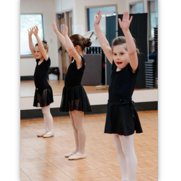 Three young girls in black leotards practicing ballet with arms raised.