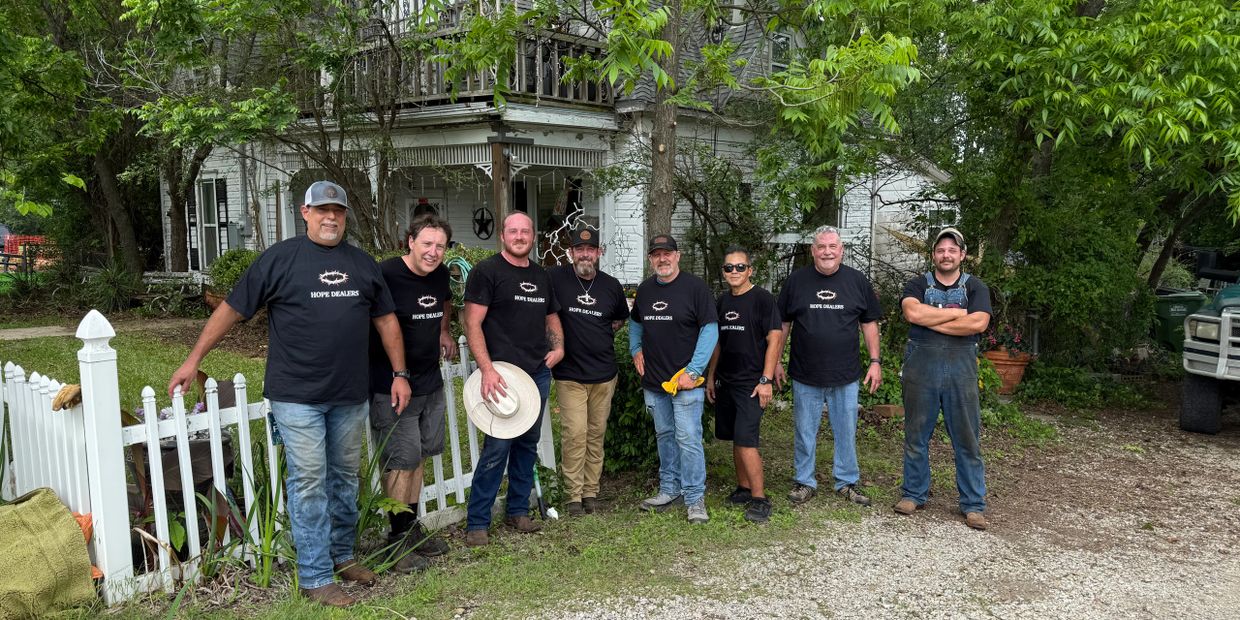 Group of eight men wearing 'Hope Dealers' shirts standing outside a house.