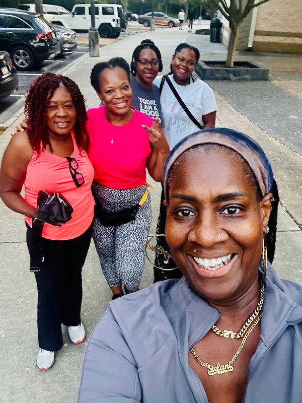 Five women smiling happily outdoors on a sidewalk.