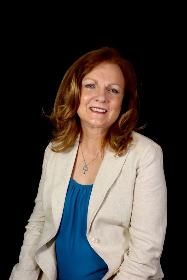Smiling woman with red hair wearing a beige jacket and blue top.