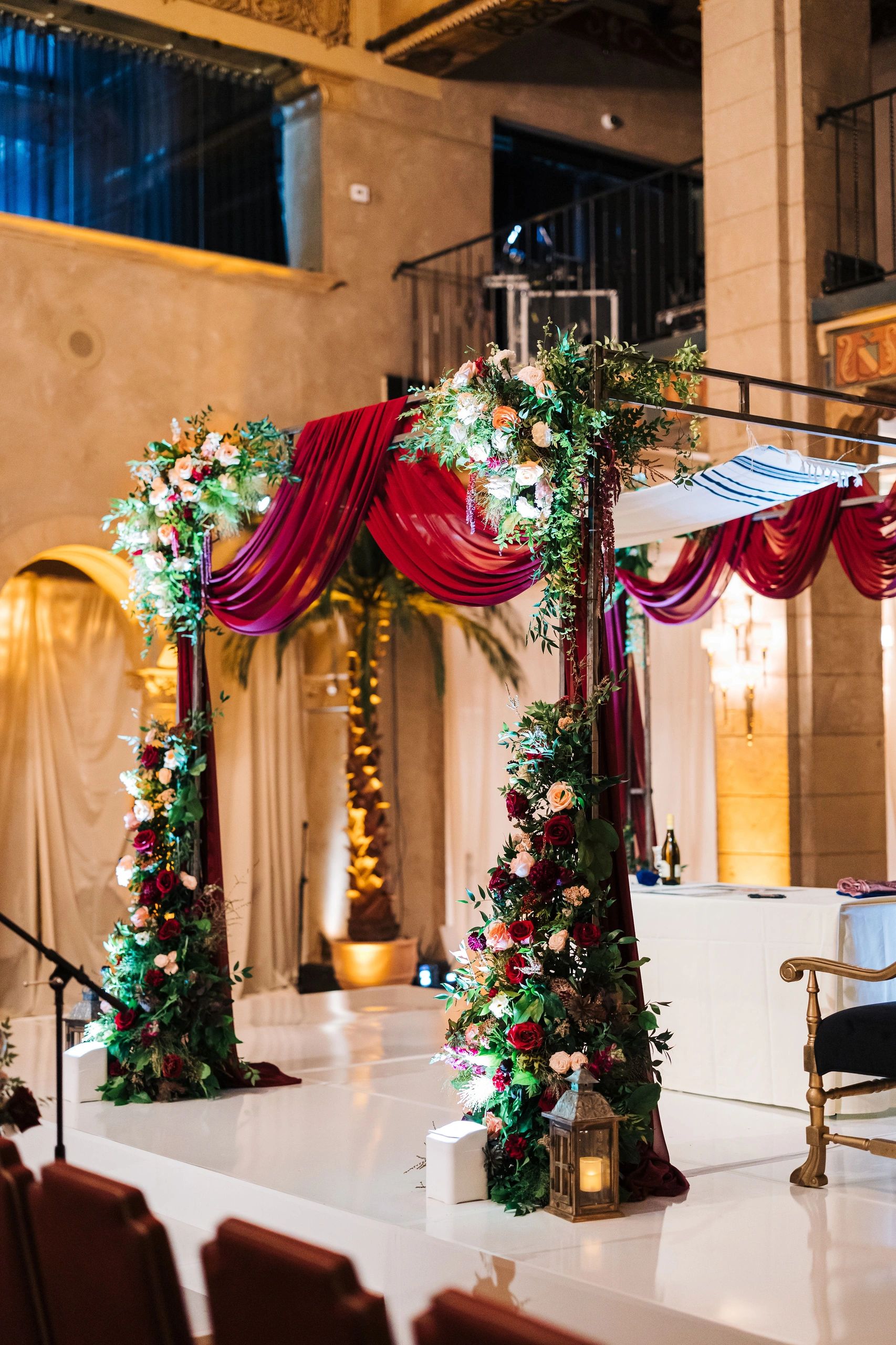 Floral-decorated wedding chuppah with burgundy drapes in an elegant venue.