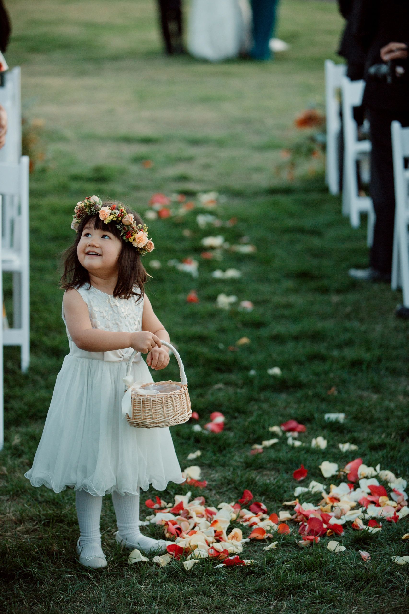 A smiling flower girl in a white dress with a basket of petals at a wedding.