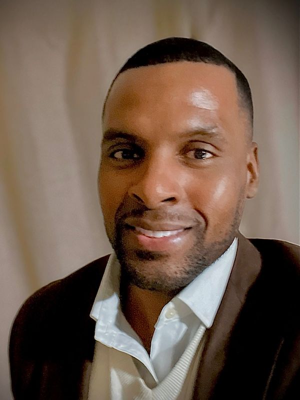 Smiling man in a white shirt and dark jacket against a beige background.