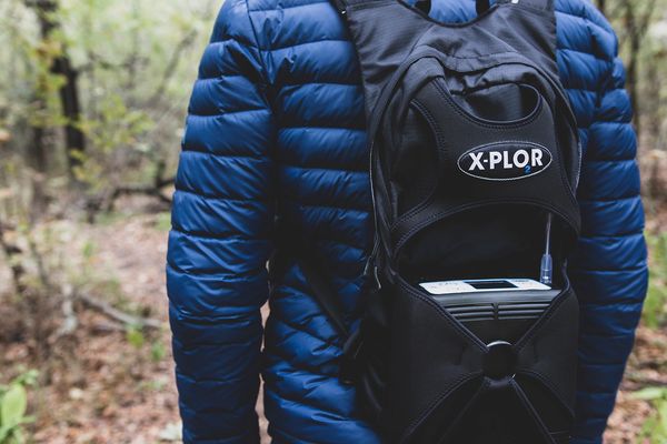 Person wearing a blue jacket with an X-PLOR backpack in the woods.