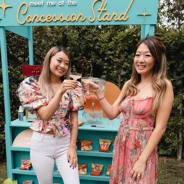 Meet me at the Concession Stand rental with two girls doing cheers with two drinks in front of stand