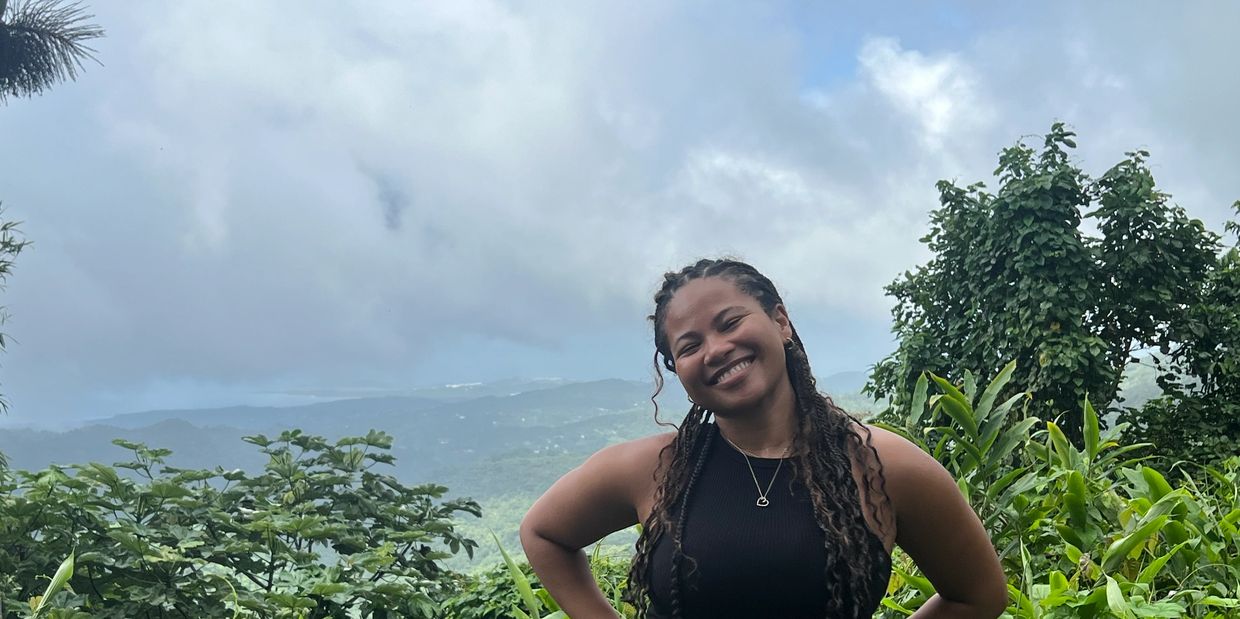 Smiling woman posing outdoors with lush greenery and cloudy mountains.