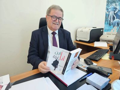 Older man in a suit reading a book at his office desk.