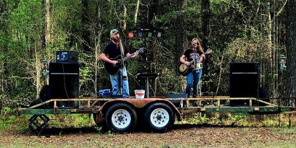Two musicians perform outdoors on a small trailer stage surrounded by trees.