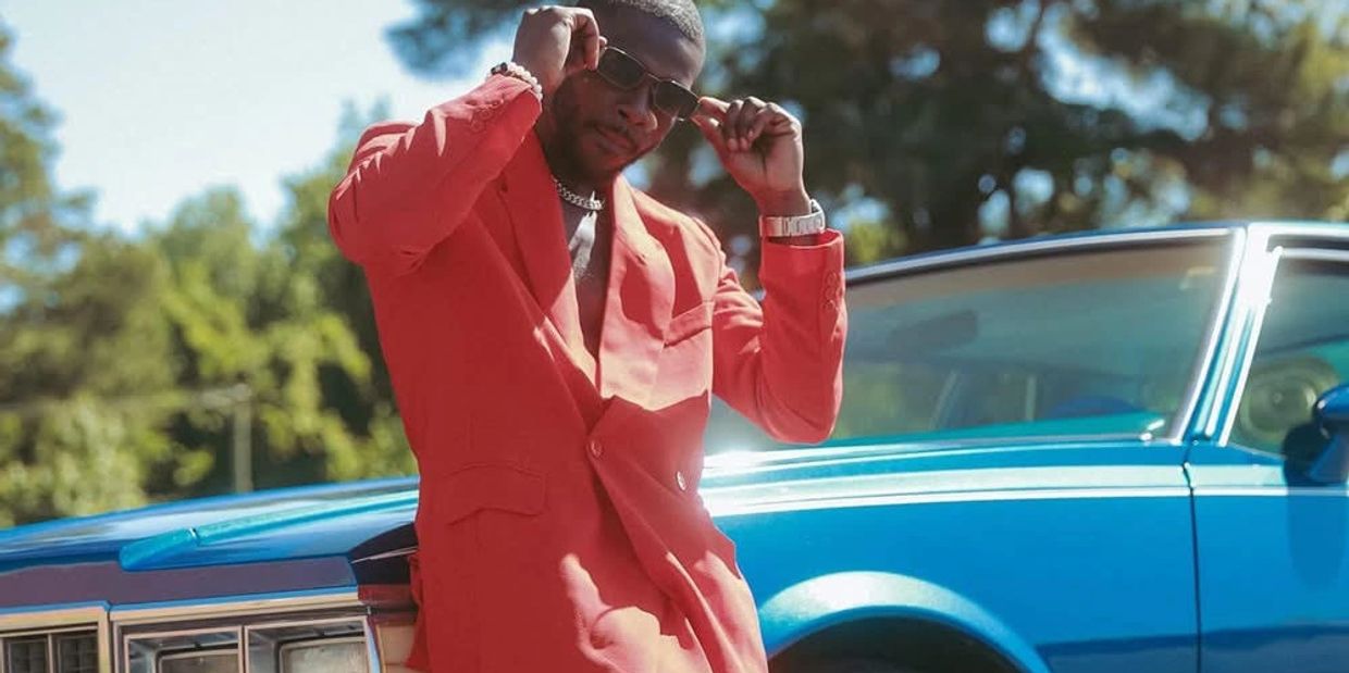 Man in red suit adjusting sunglasses beside a blue vintage car.