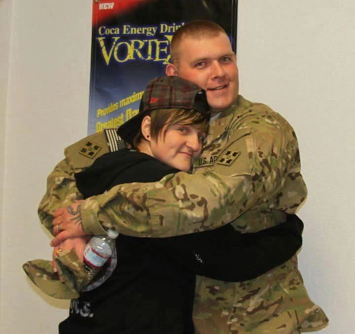 A soldier in uniform warmly hugging a young person in casual clothes indoors.