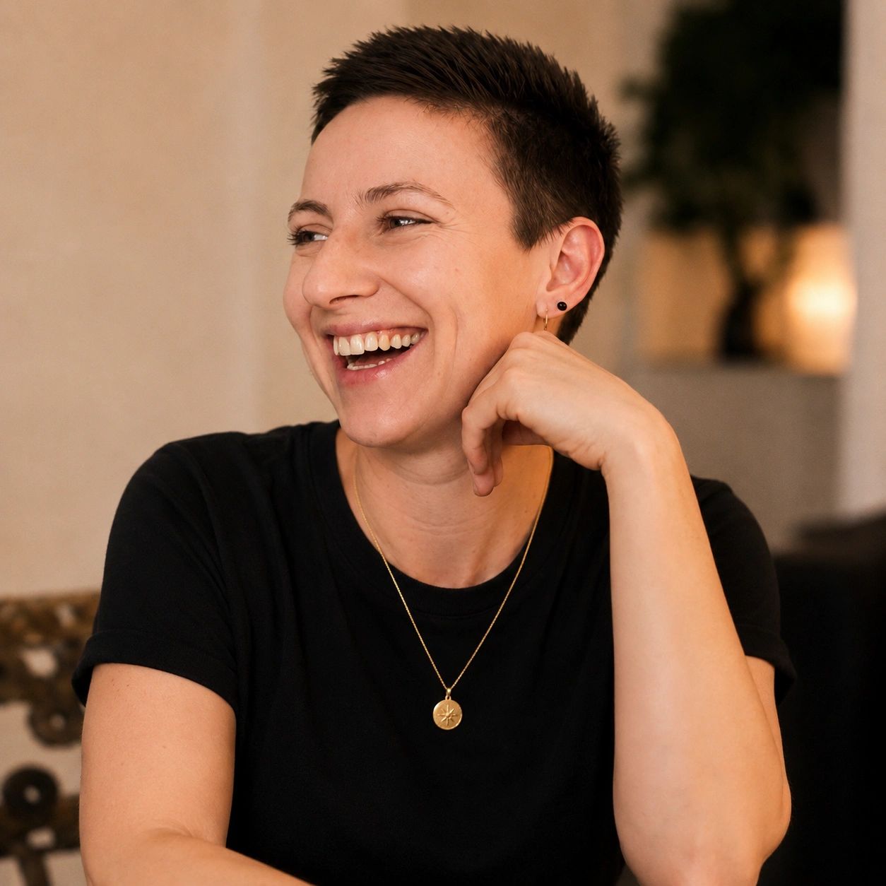 Smiling person with short hair wearing a black shirt and gold necklace.