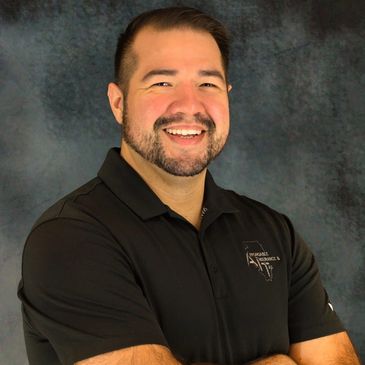 Smiling man with beard in black polo shirt standing confidently with arms crossed.