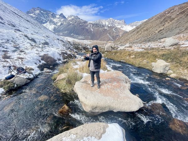 A person playing flute on a rock in a snowy mountainous stream.