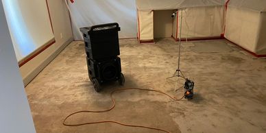 Room with covered furniture and drying equipment on a bare floor.