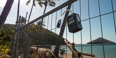 A black device attached to a metal fence by a beach with palm trees and an island in the background.
