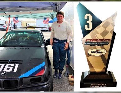 Man in racing suit next to a BMW car and a third-place trophy.