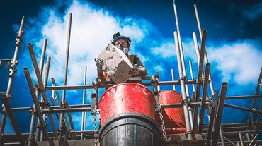 Construction worker handling cement block on scaffolding under blue sky.