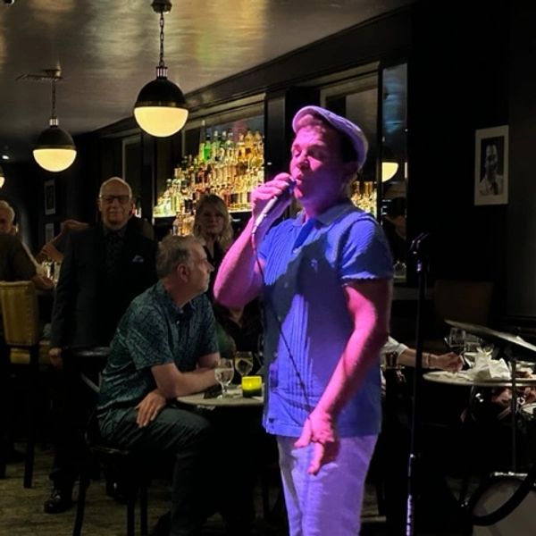 A man sings into a microphone at a dimly lit bar with an attentive audience.