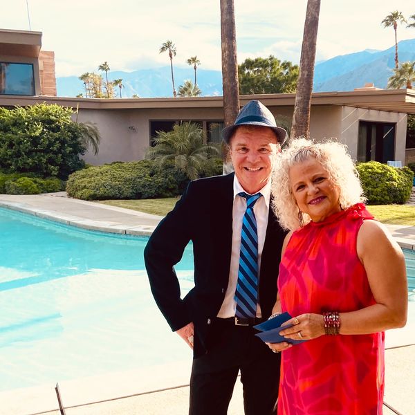 A smiling couple dressed elegantly by a pool with palm trees and mountains in the background.