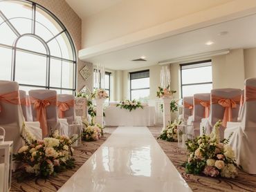 Elegant indoor wedding ceremony setup with floral decorations and peach chair sashes.