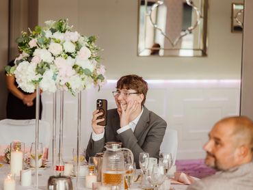 A young man in a suit smiles while video calling on his phone at a decorated event table.
