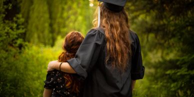 Graduate in cap embraces a child in a green forest.