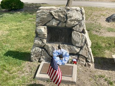 Memorial stone with plaques, a wreath, and an American flag in a grassy area.