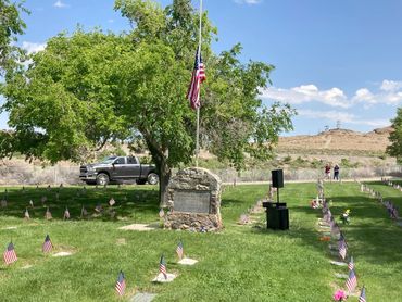 Memorial with American flags and a stone marker under a tree on a sunny day.