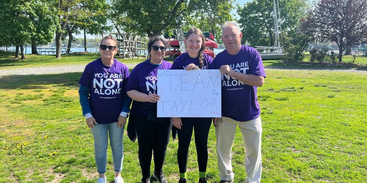 Four people in purple shirts hold a Team Taylor sign outdoors on a sunny day.