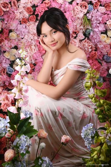 Young woman in a floral dress poses gracefully against a vibrant flower wall.