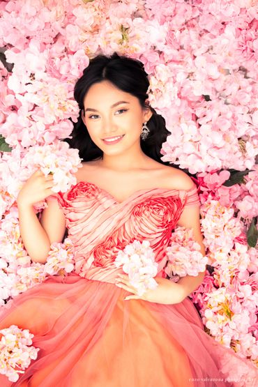 A woman in a peach gown lies amidst pink flowers, smiling gently.
