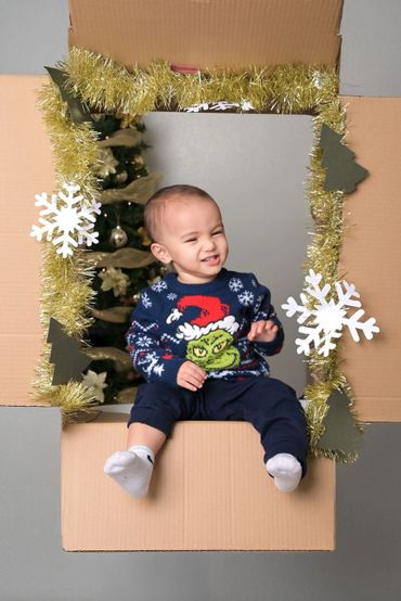 Child in festive sweater sitting in decorated cardboard frame with Christmas tree in background.