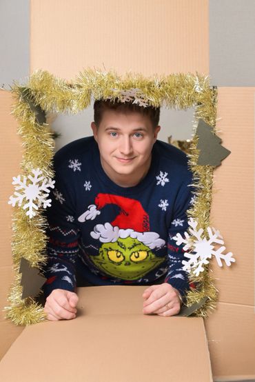 Man in a festive Grinch sweater peeks through a decorated cardboard frame.