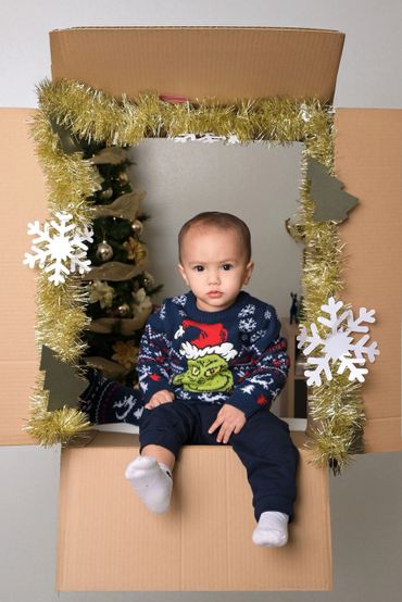 Toddler in a festive sweater sitting inside a decorated cardboard frame.
