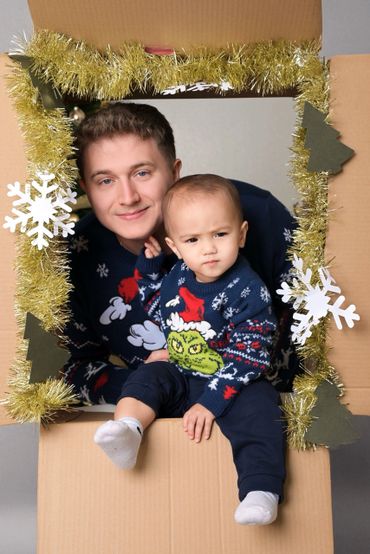 Father and son in matching holiday sweaters pose inside decorated cardboard frame.
