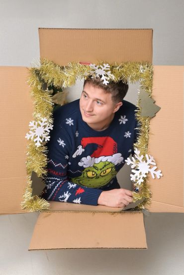 Man in a Grinch sweater peeks out of a decorated cardboard box.