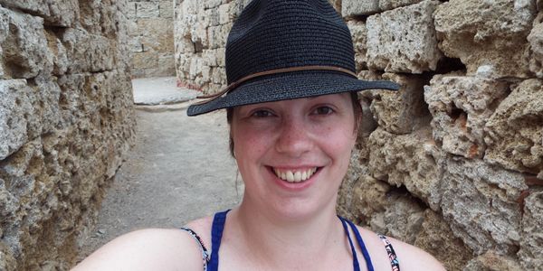 Leigh smiling at a camera, wearing a black hat and blue top, in an ancient brick walled alley.