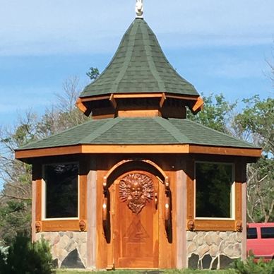 An octagonal wooden gazebo with a green roof and ornate door featuring a sun design.