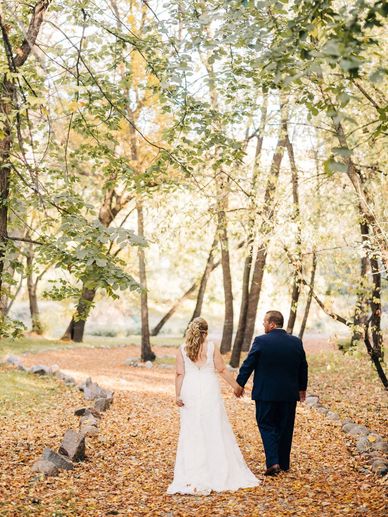 A bride and groom walking hand in hand through a sunlit forest with autumn leaves.
