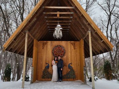 Couple embracing in front of a wood chapel with peacocks and a chandelier, in a snowy setting.