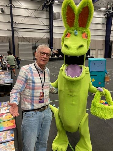 An elderly man poses with a person in a colorful dinosaur costume at a convention.