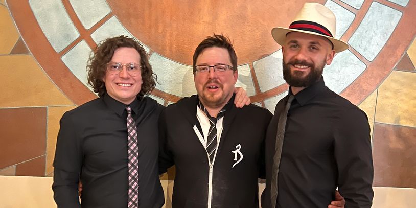 Three men smiling, dressed in black with ties, one wearing a white hat.