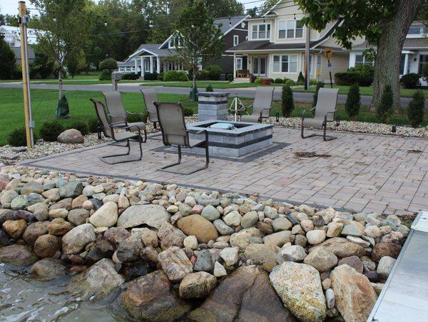 Outdoor patio with chairs around a fire pit near a rocky water edge.