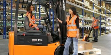 Two women in safety vests working with a forklift in a warehouse.